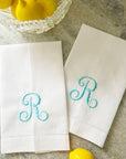 Two white linen guest towels with turquoise monogram 'R' on a stone surface with lemons.
