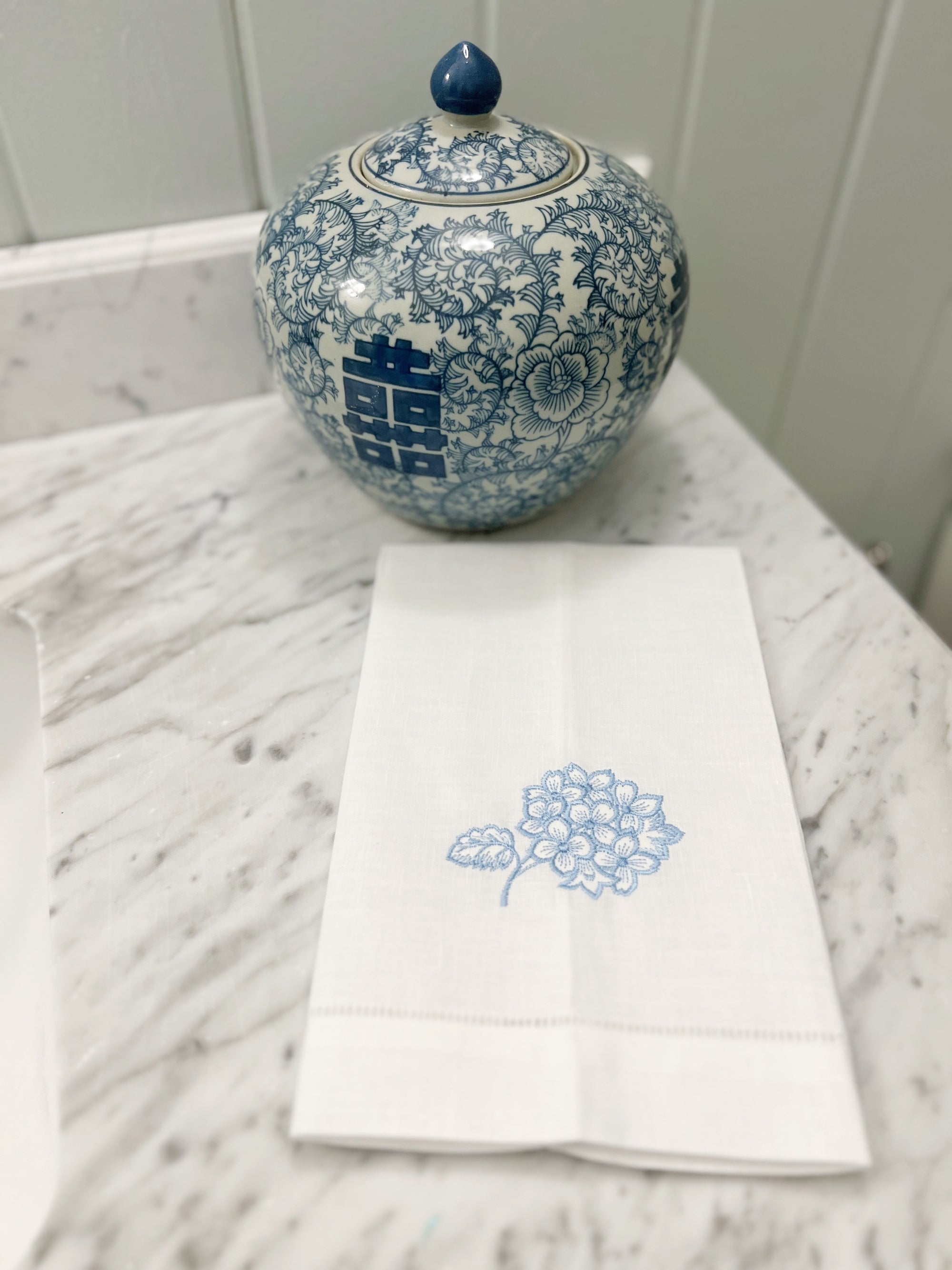 white guest towel with embroidered blue hydrangea with blue and white chinoiserie jar on a marble bathroom counter