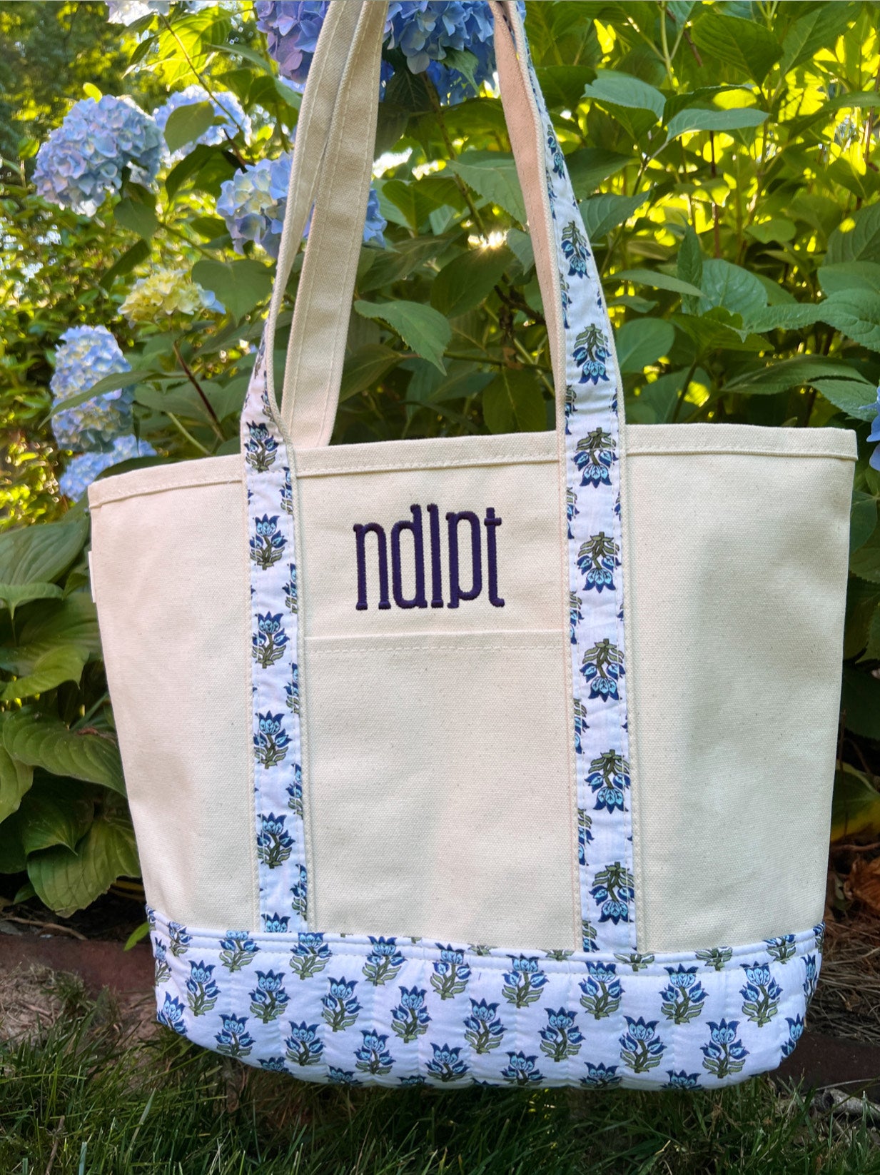 Canvas tote bag with block print accents and 'ndipt' monogram against a green leafy background