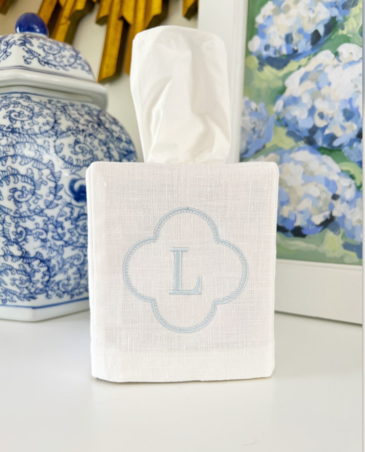 White tissue box cover with blue monogram 'L' on a surface with decorative items in the background.