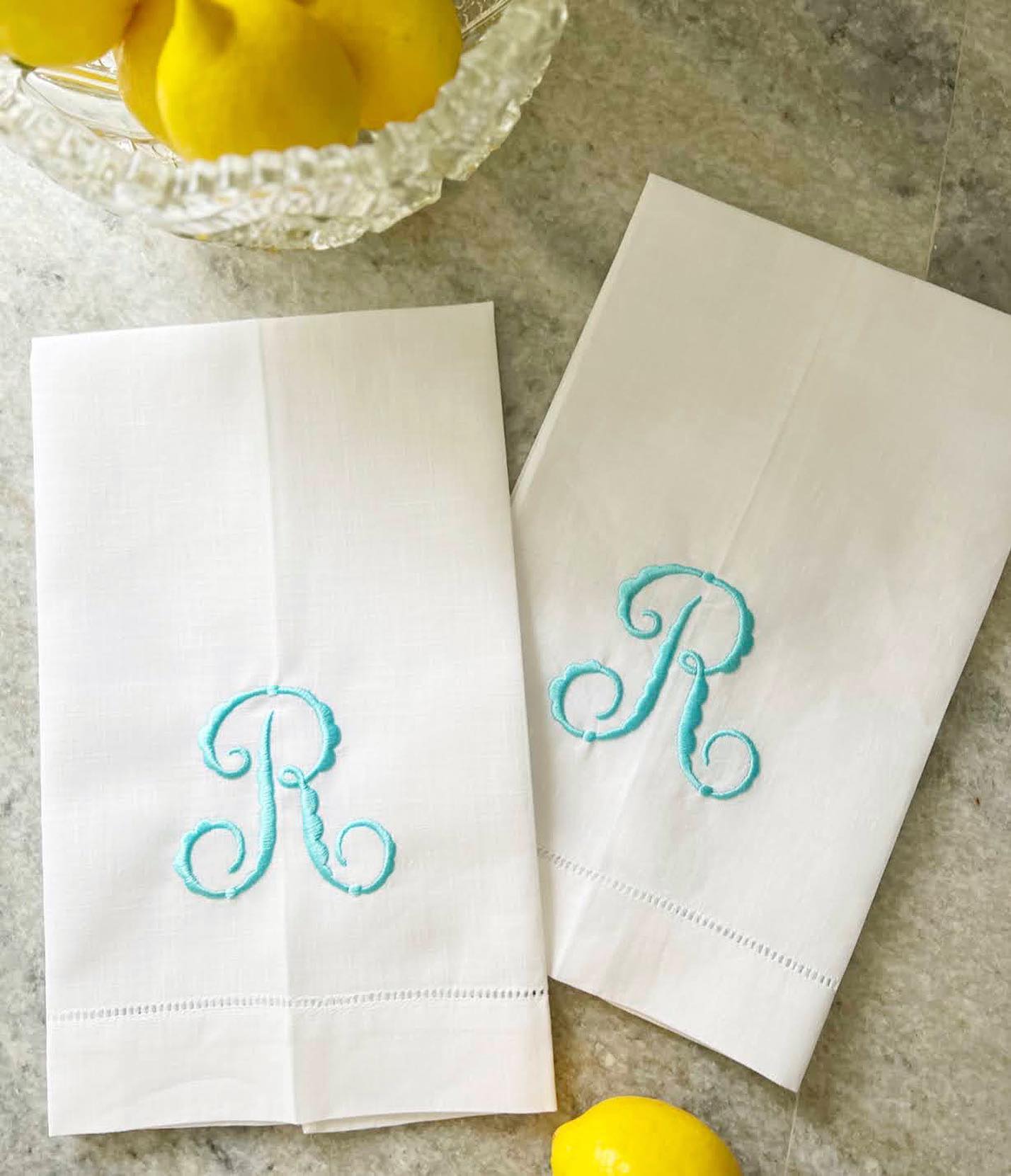 Two white linen guest towels with turquoise monogram 'R' on a stone surface with lemons.